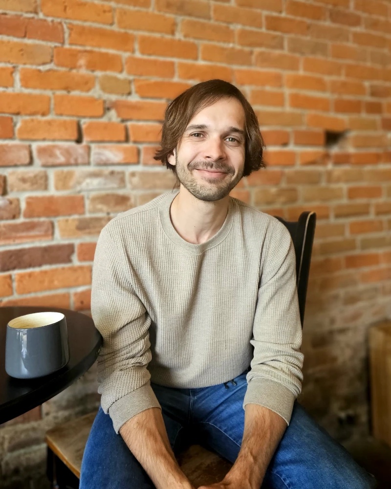 Nic Kozushko smiling, seated in front of a brick wall